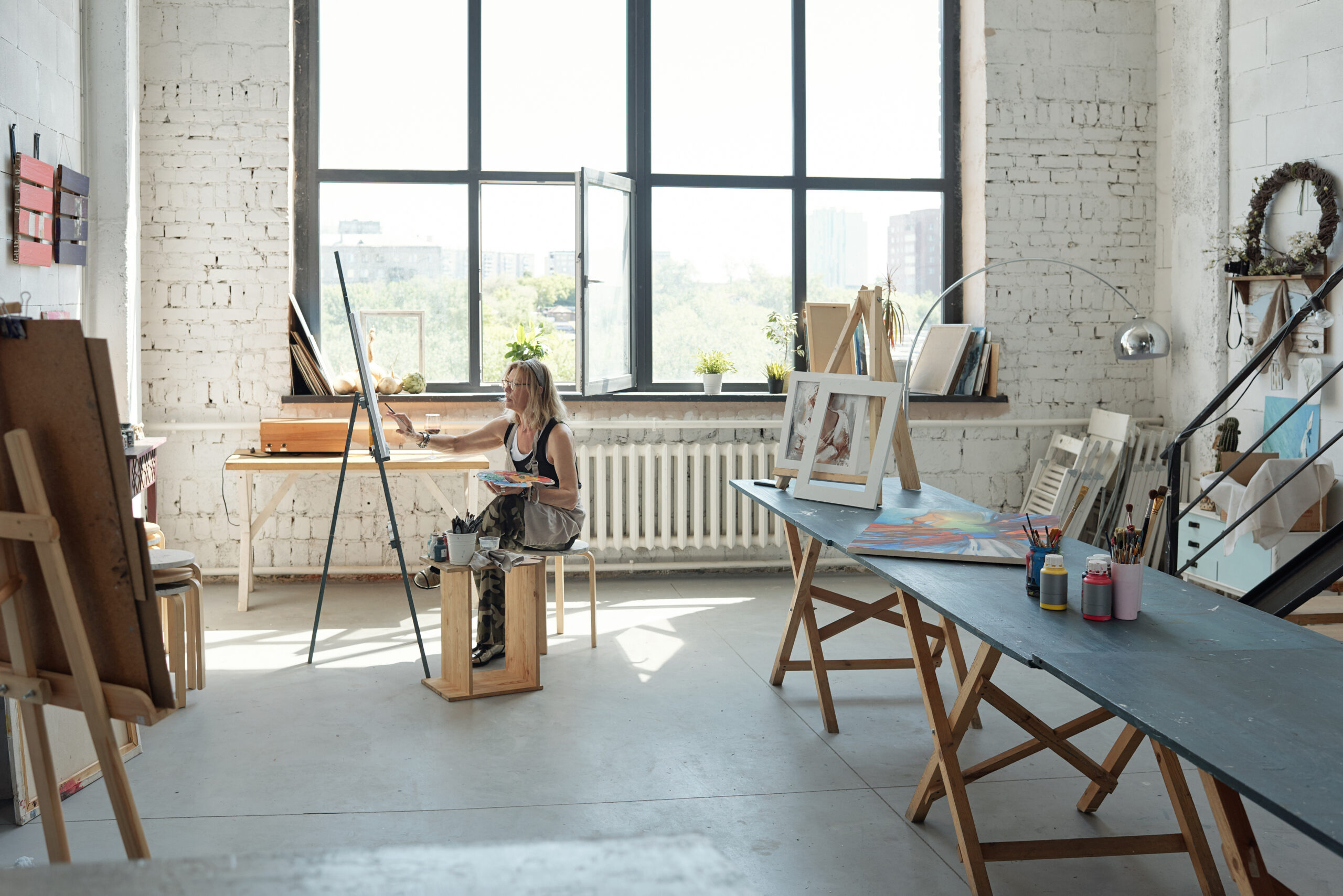 Artista feminina sentada em frente à tela e pintando em um estúdio pessoal espaçoso e iluminado