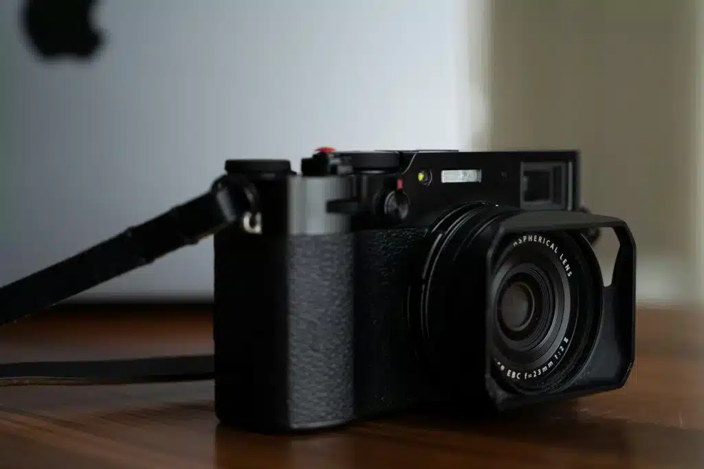 Câmera sem espelho Fujifilm x100v preta sobre uma mesa com um MacBook Pro ao fundo