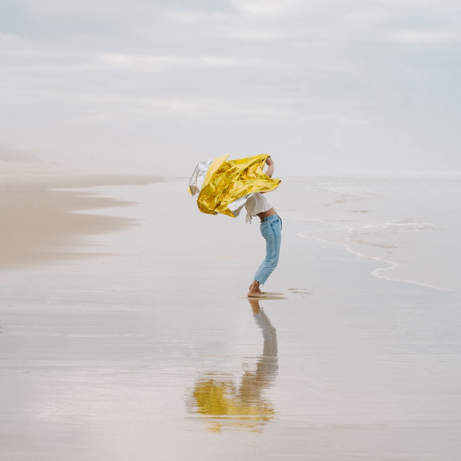 woman in jeans a tshirt and bare feet standing in the wet sand at a cloudy beach, shaking bright yellow fabric. titled "New summers" by Émilie Möri