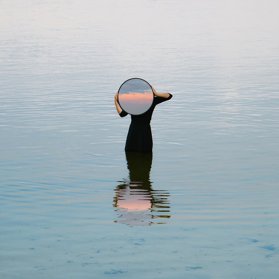 woman standing is water wearing a black dress and holding a round mirror that reflects the sunset. Titled "The shades of my lake" by Émilie Möri