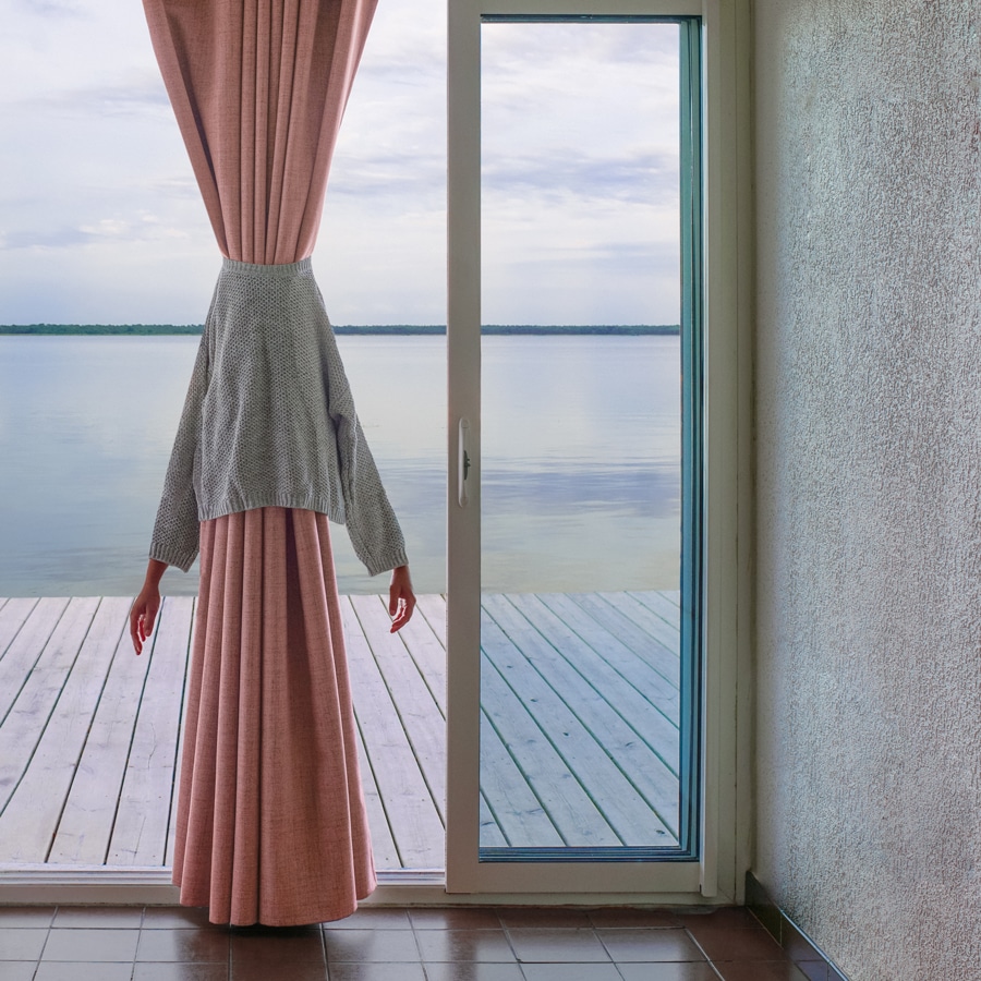 view from inside a room with glass sliding doors, looking out at a deck over glassy water. A gray sweater is around a long pink curtain, a woman's hands emerging from the sleeves of the sweater. Image by Émilie Möri