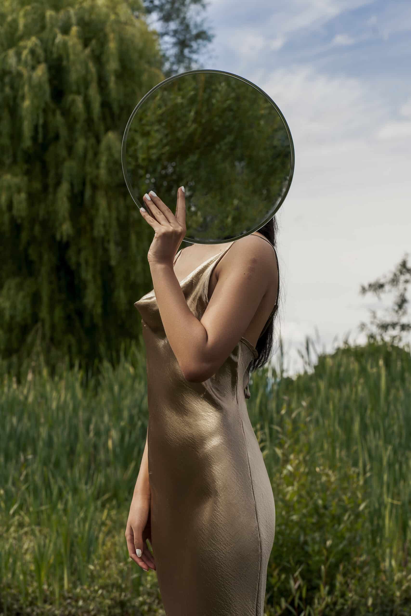 Woman wearing fitted bronze dress holding a circular mirror in front of her face, reflecting the green field and trees around her. Photo by Jada Jimpinu