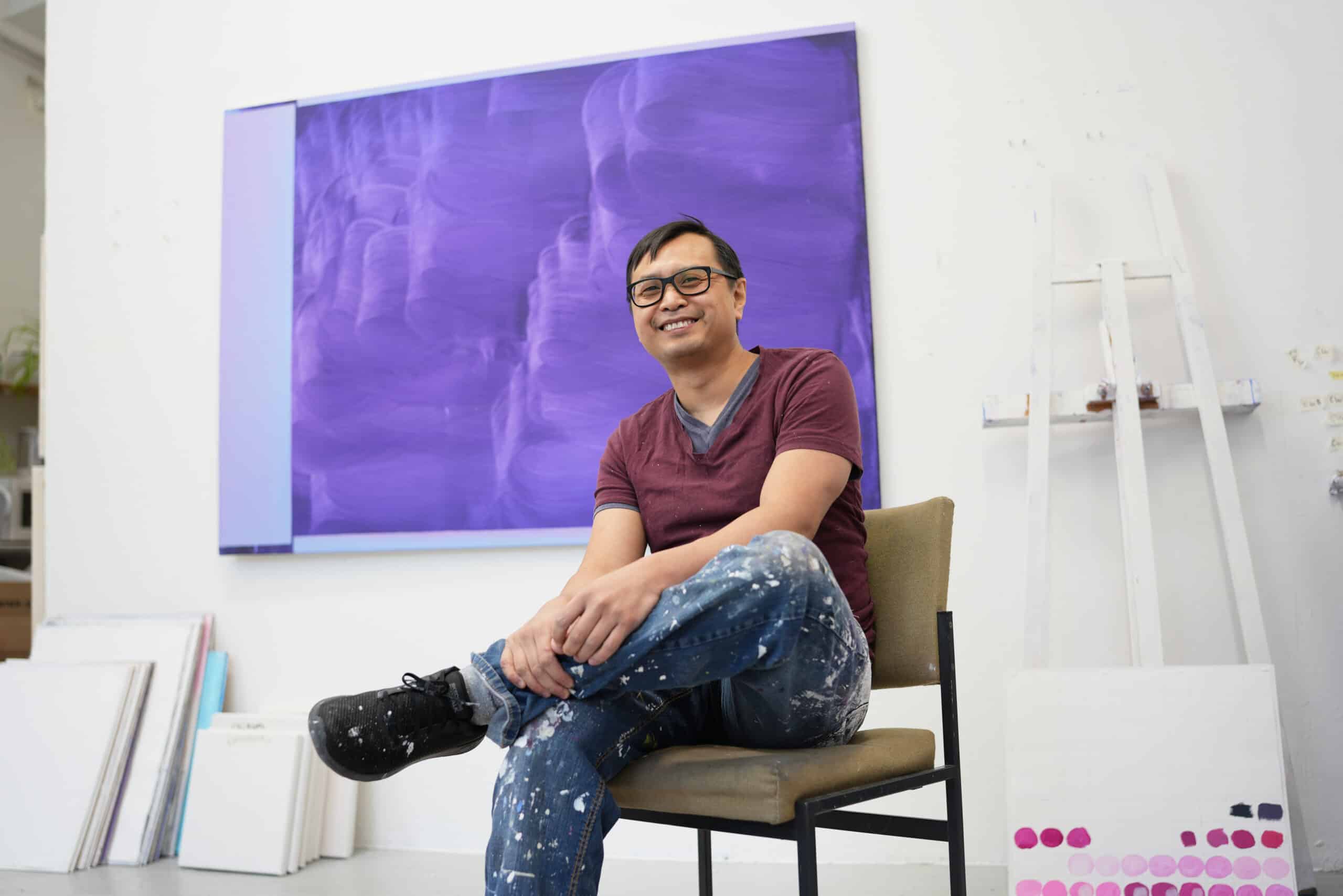 Retrato de Nam Duc Nguyen sonriendo y mirando a la cámara, sentado en una silla frente a un cuadro morado con grandes marcas de pincel en su estudio.