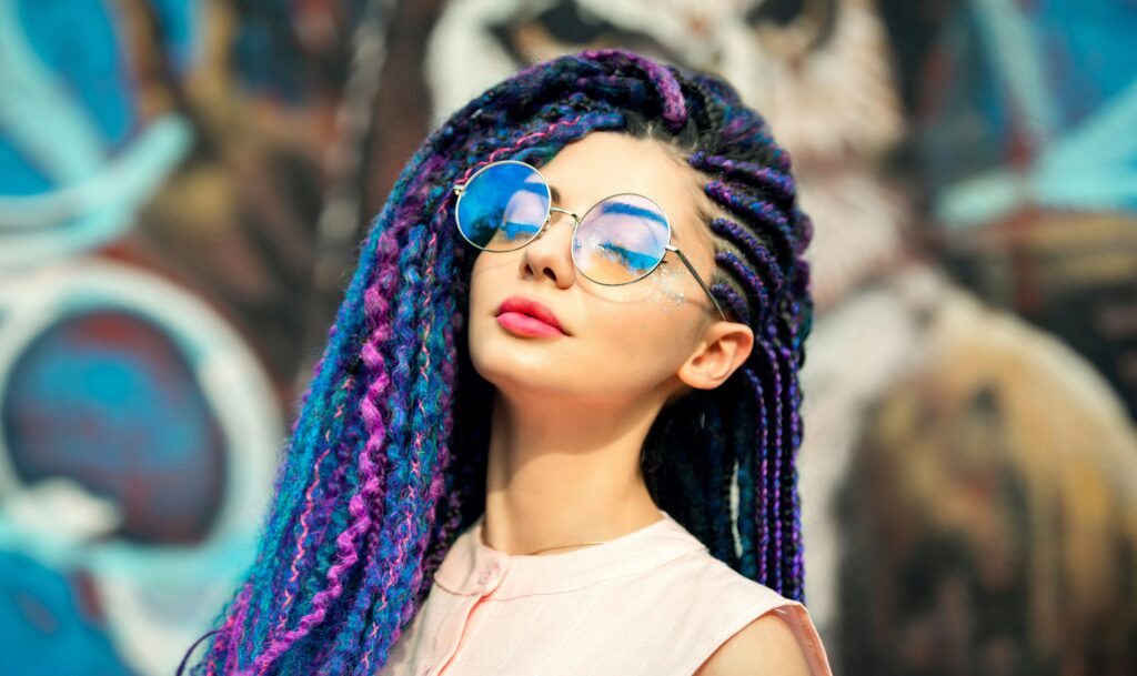 retrato de mujer a la luz del sol con trenzas de colores, pintalabios rojo y gafas de sol traslúcidas con los tonos del arco iris, con la cara inclinada hacia arriba