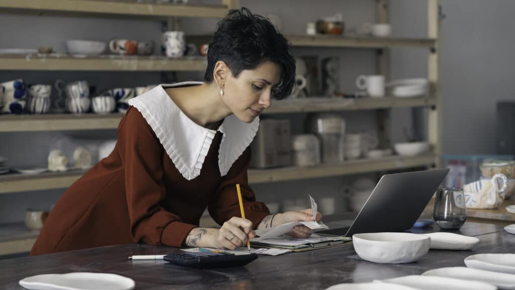 woman in ceramic studio filing taxes with laptop and receipts