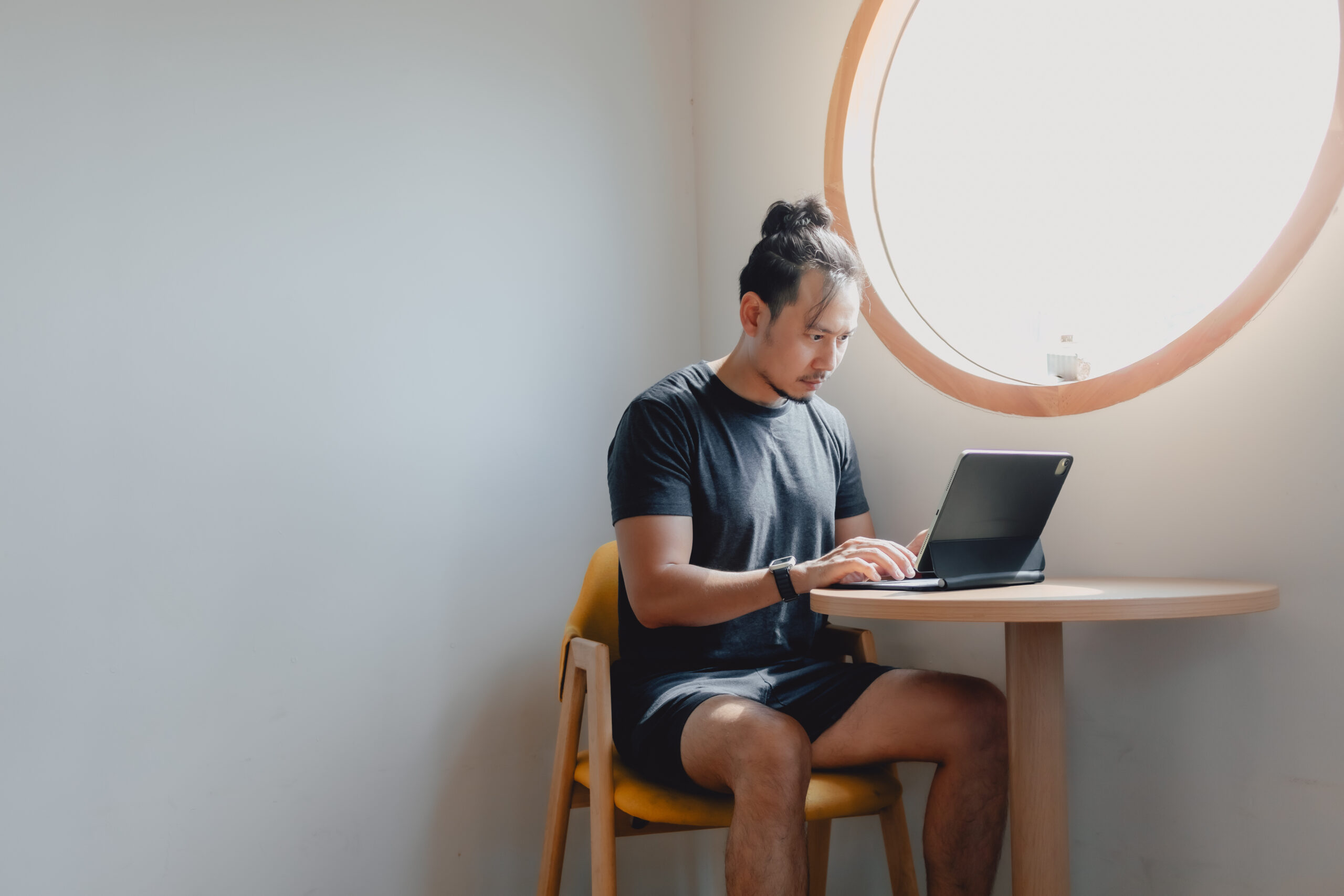 Freelance man working on computer tablet at small table by round window.