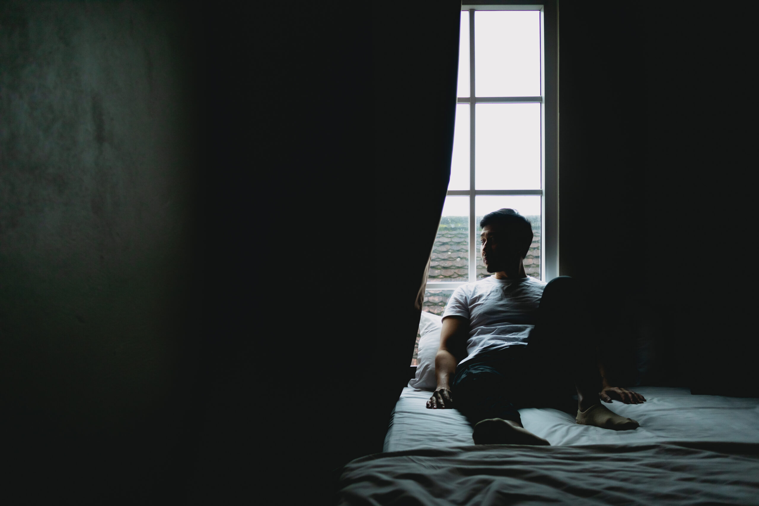 young man sitting in bed alone in dark bedroom with light from curtain