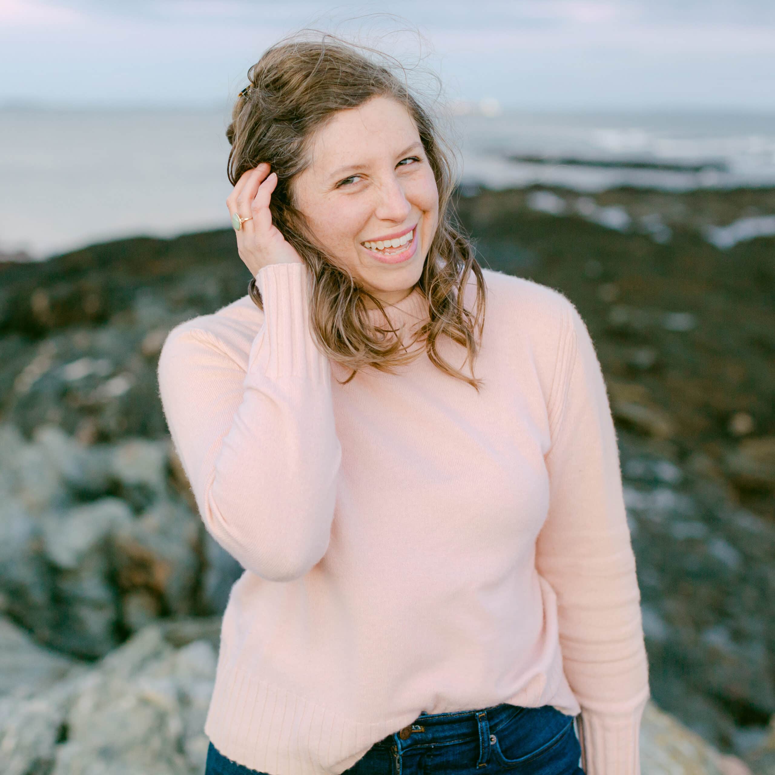 portrait d'une femme aux cheveux bruns portant un chandail rose pâle sur une plage rocheuse