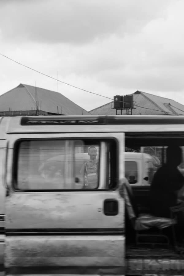 scène de rue en noir et blanc montrant un garçon à travers les vitres d'une camionnette, avec des maisons en arrière-plan