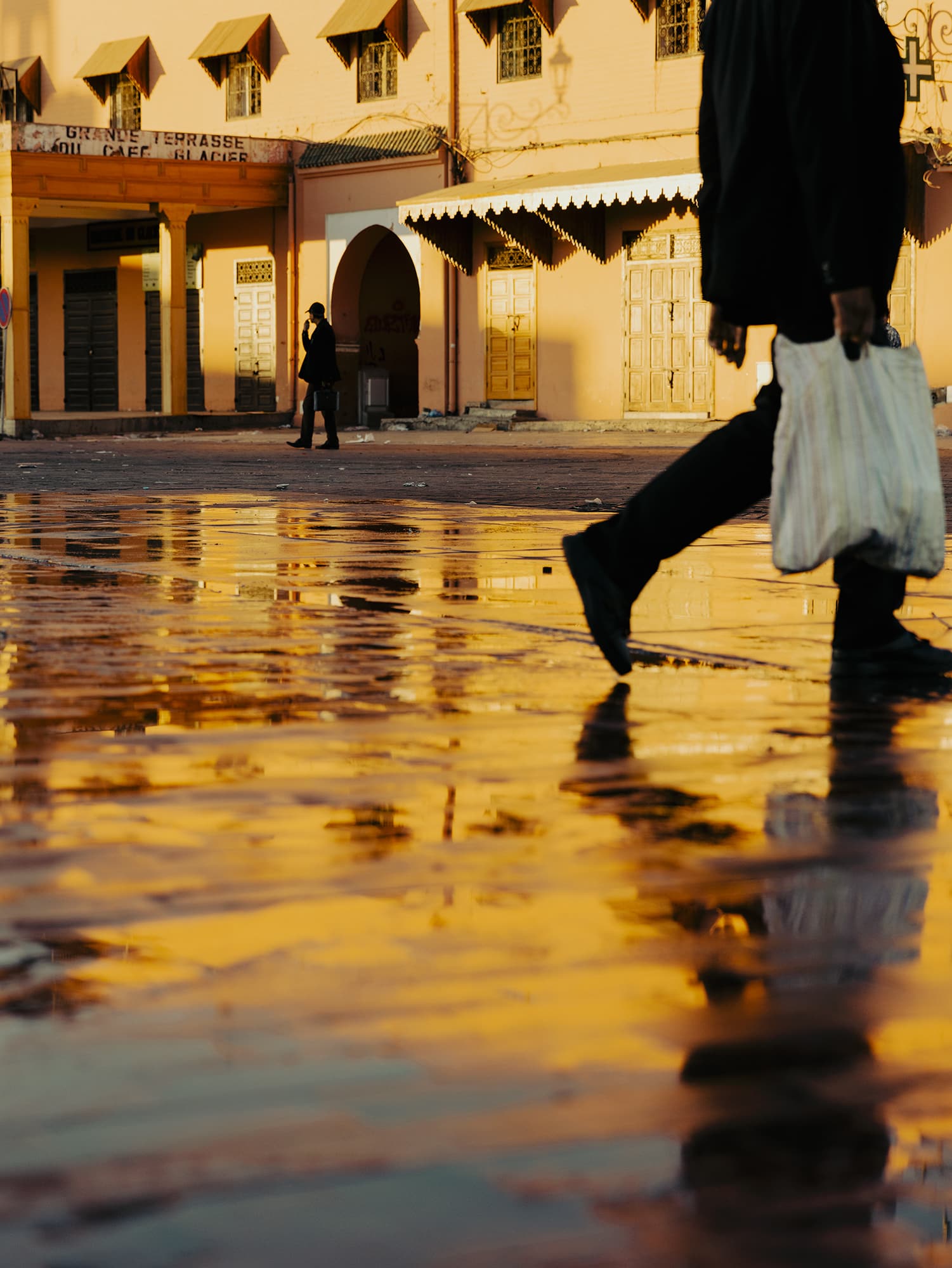 low angle street photograph after a rainstorm, with the golden glow of a building in the background reflecting in the shallow puddles on the pavement. Photo by Grant Harder