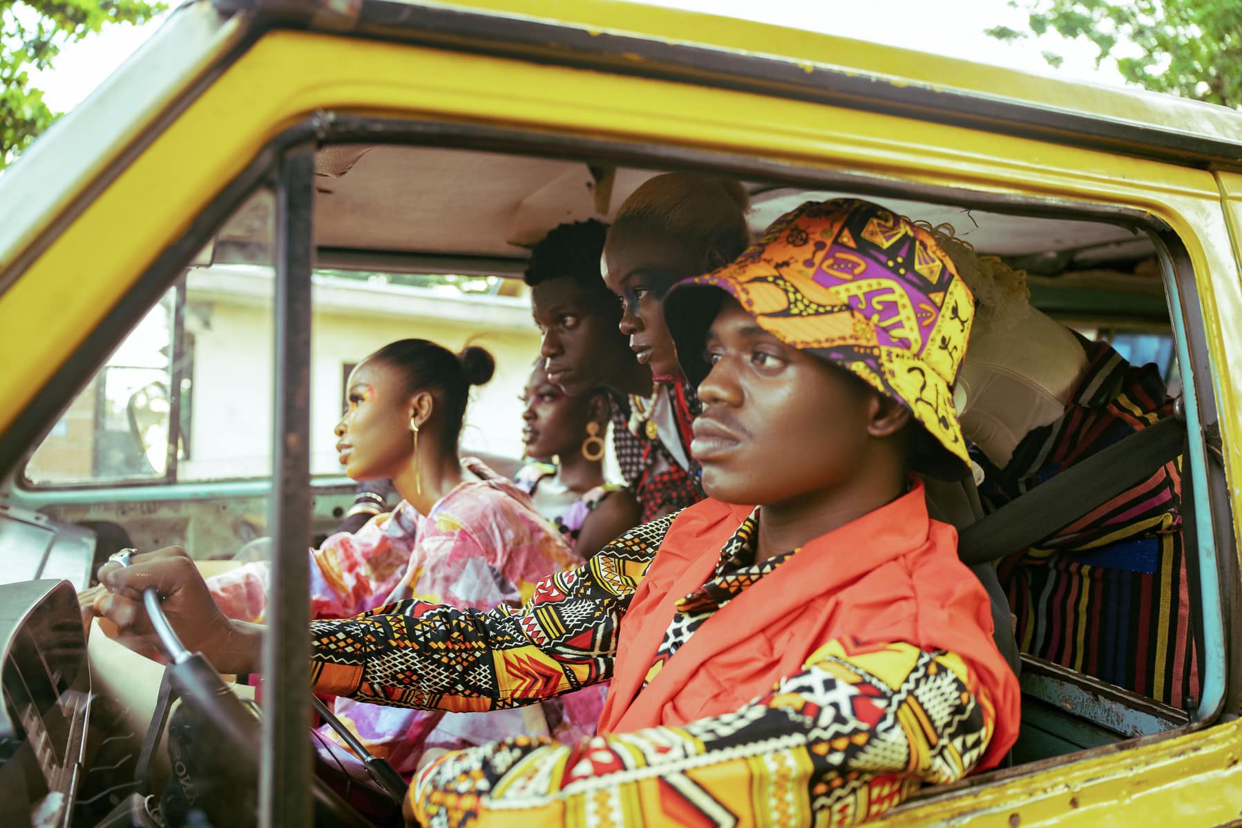 five people piled into a yellow van and leaning toward the font windshield. From the Japa Syndrome photo project by Jada Jimpinu