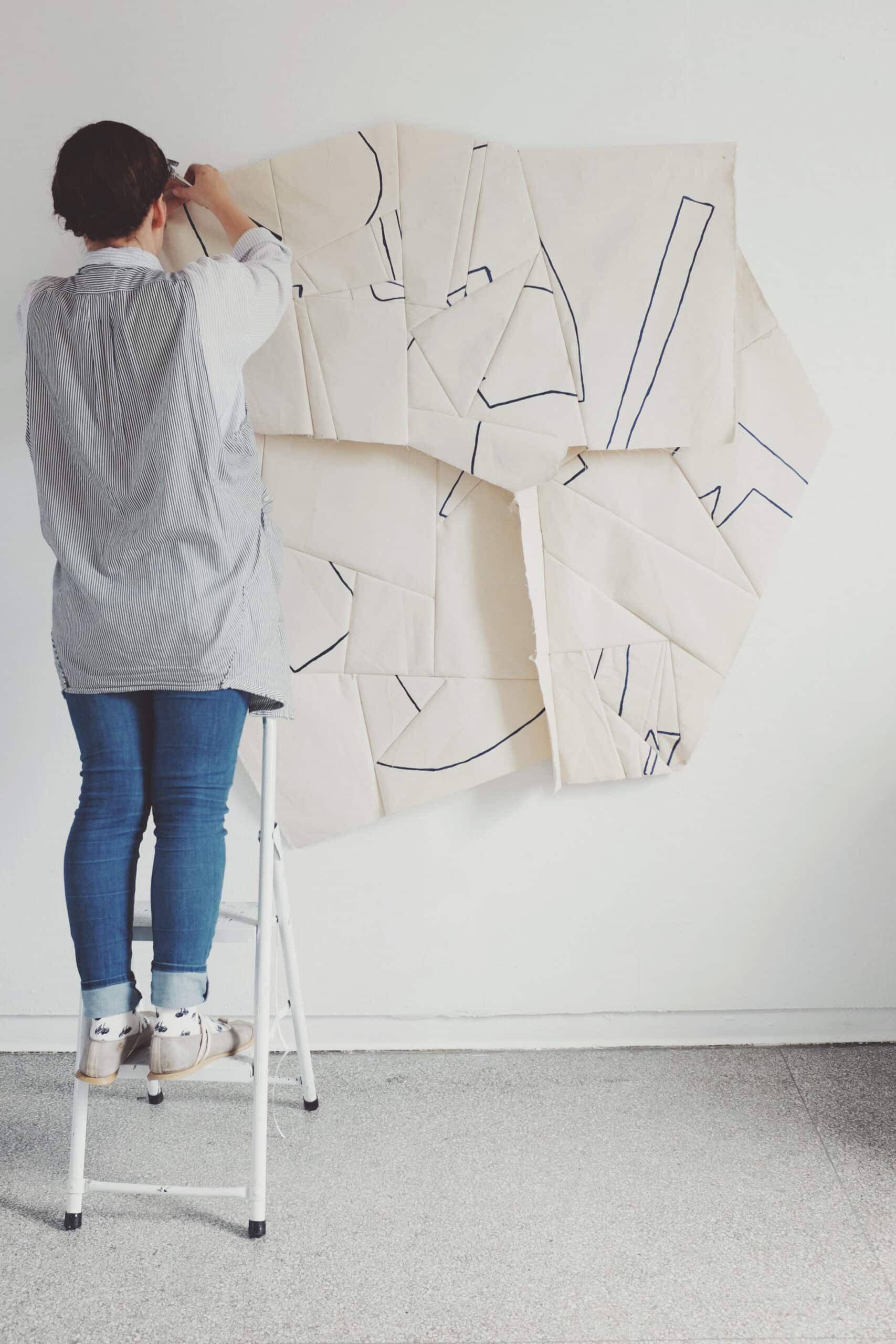 image of a woman standing on a stepstool hanging artwork on a white wall. Processed with VSCOcam with f2 preset