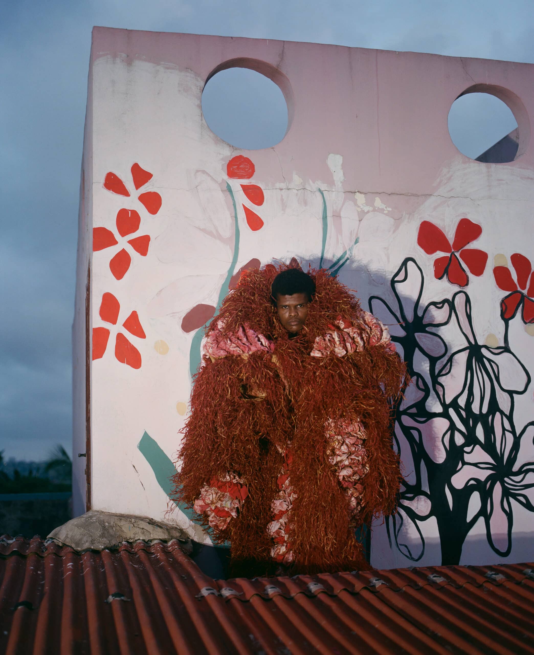 man swathed in garment made from fabric and rust colored dried grasses posed in front of a floral mural on a red rooftop. title: Untitled (Rooftop) photograph by Delali Cofie