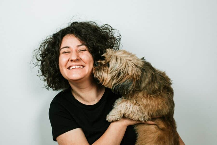 curly haired woman laughing and holding her dog near her face