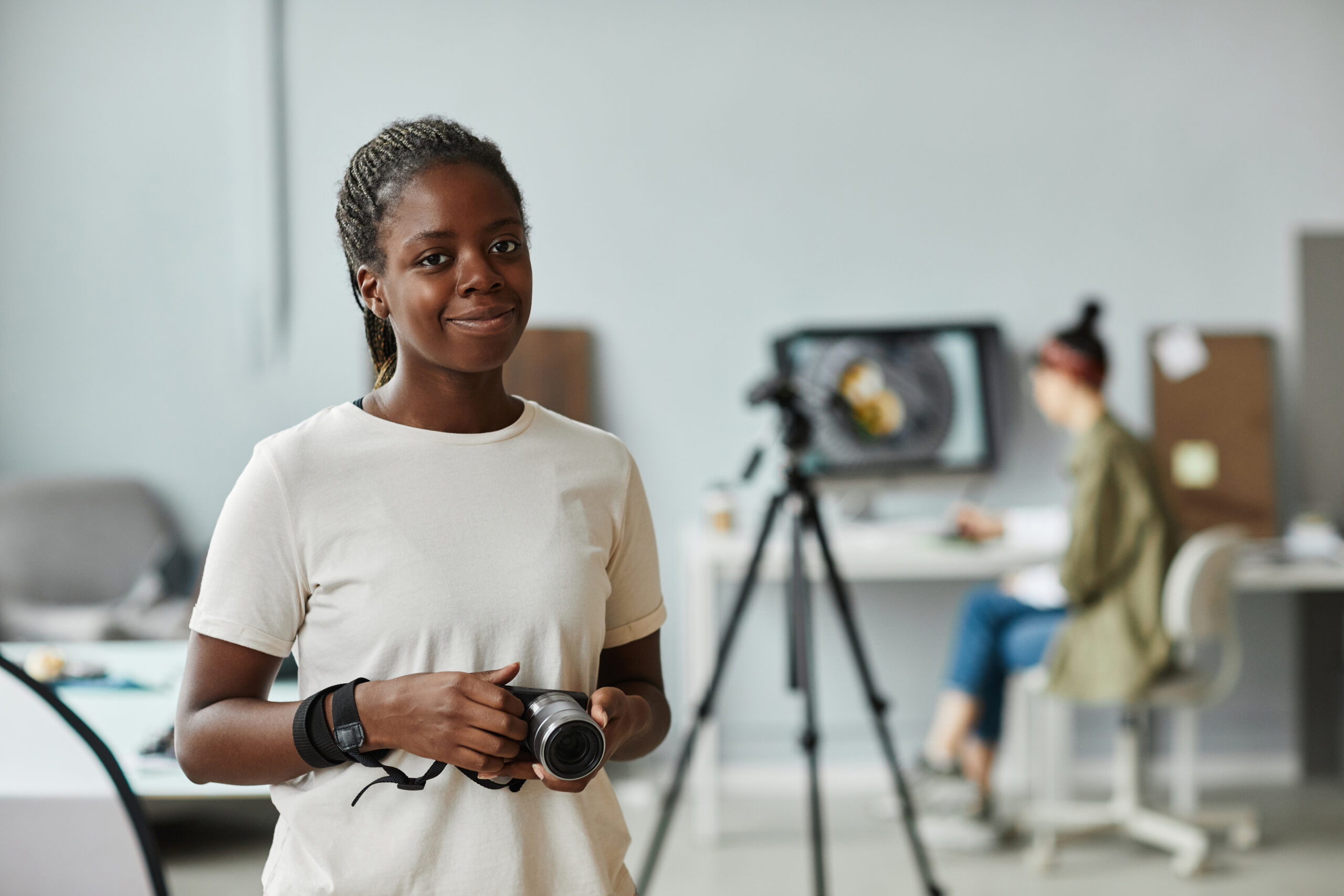 Portrait à la taille d'une jeune femme afro-américaine souriant à l'appareil photo, debout dans un studio photo, avec en arrière-plan une personne effectuant des retouches sur un ordinateur de bureau.