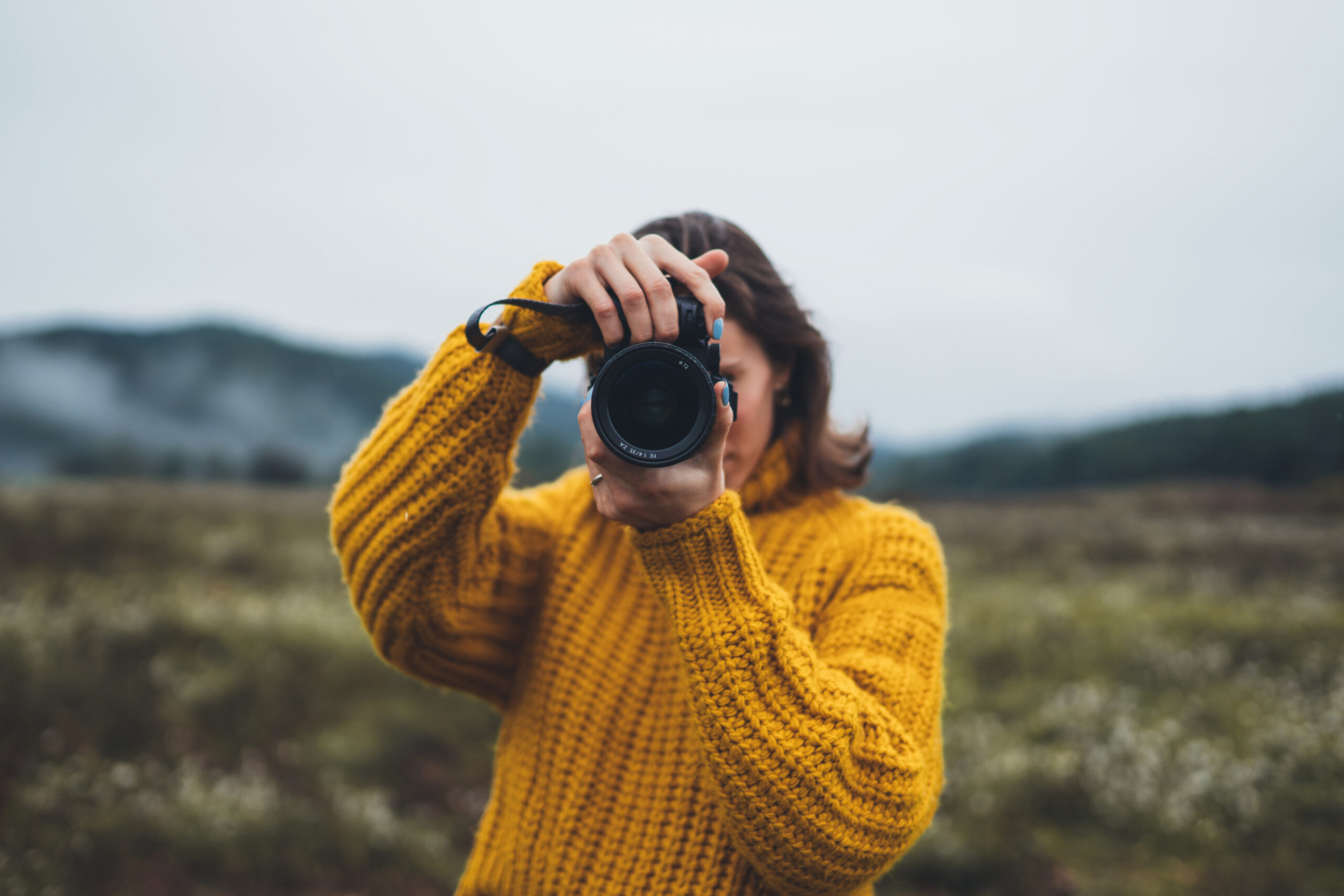 female photographer in yellow sweater pointing camera at viewer in autumn on a foggy mountain