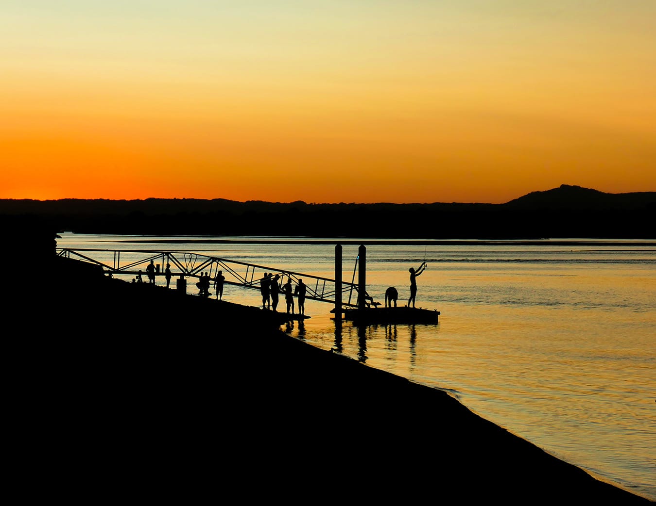 silhouette image of people fishing off the end of a dock in the distance during sunset